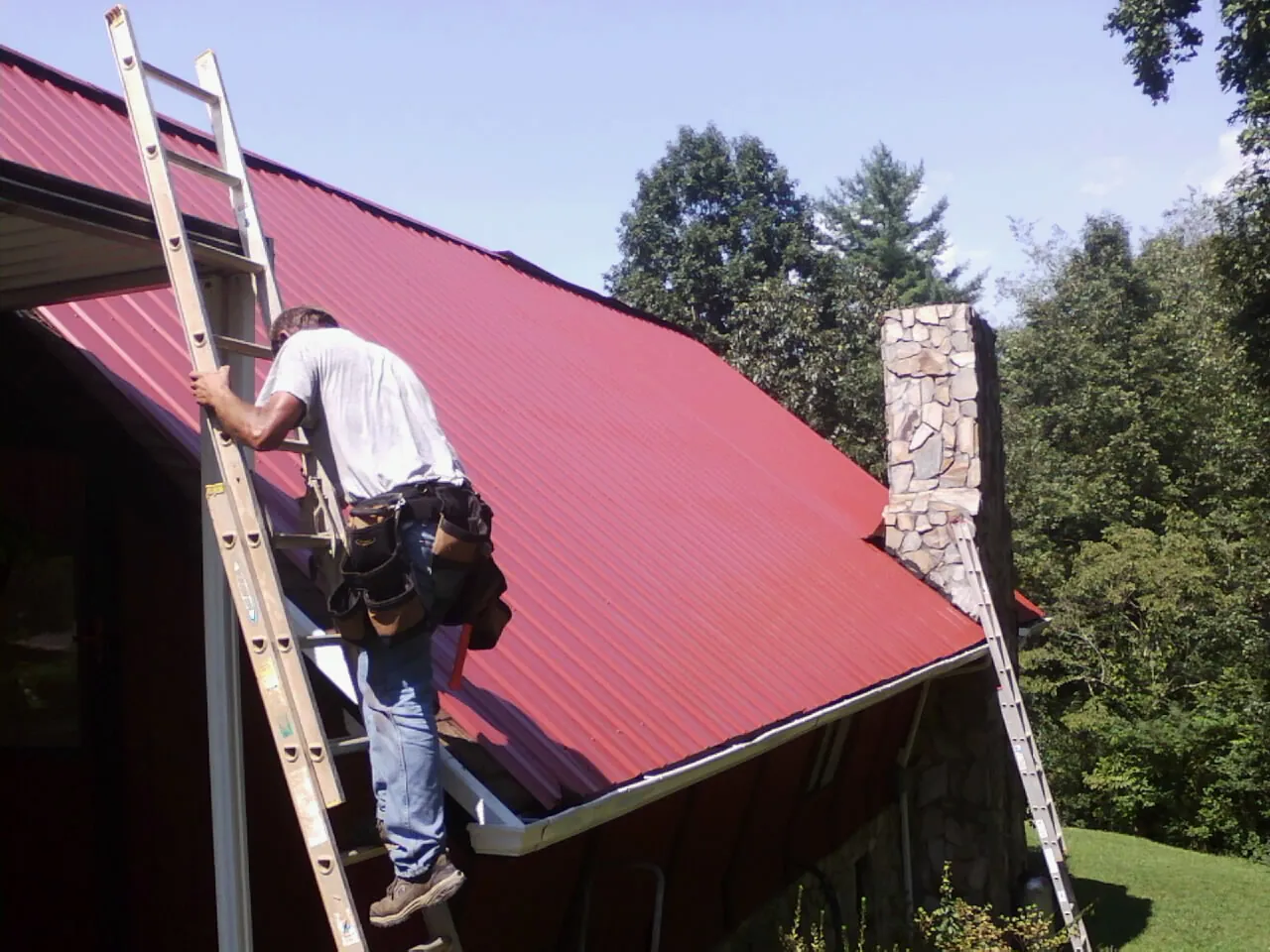 Roofer on a ladder beside a red metal roof and stone chimney — Soffit & Fascia Repair by Roofing Brothers in Lisle